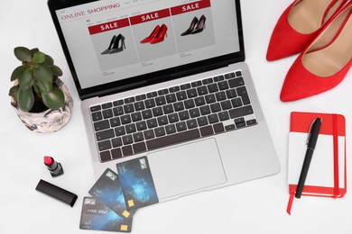 Photo of Online store website on laptop screen. Computer, credit cards, women's shoes, stationery and lipstick on white background, above view