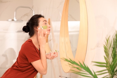 Photo of Young woman applying mask on her face near mirror in bathroom