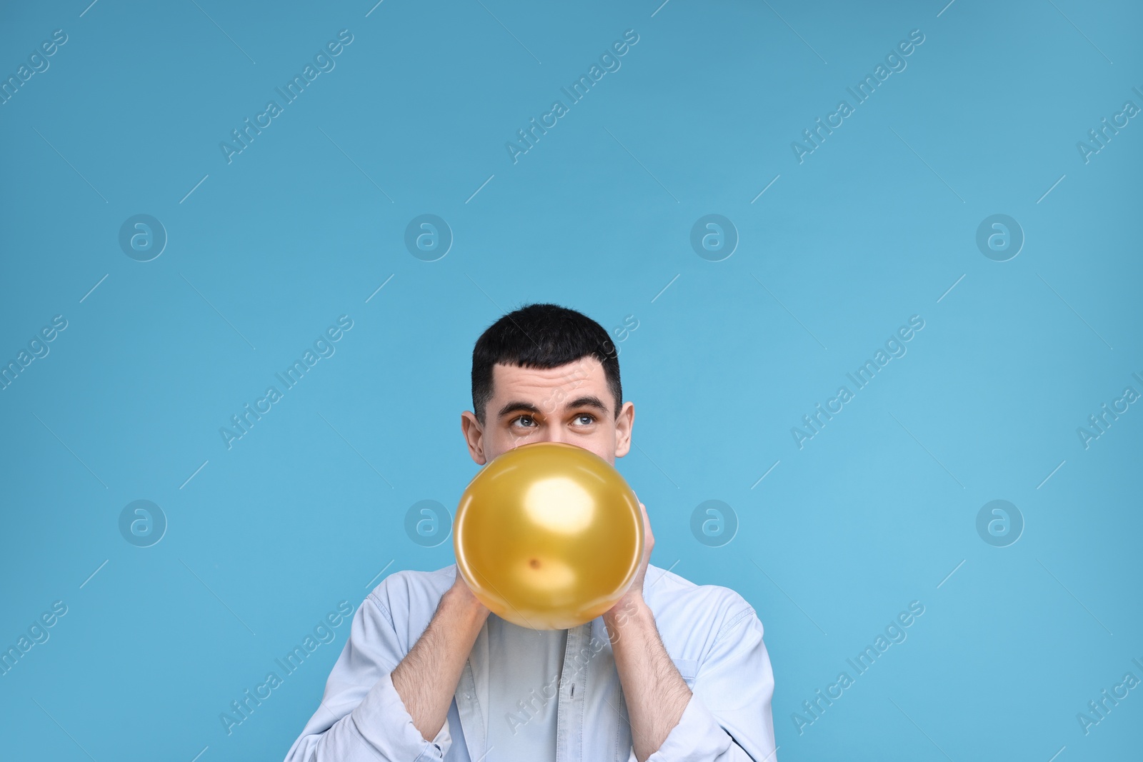 Photo of Young man inflating golden balloon on light blue background. Space for text