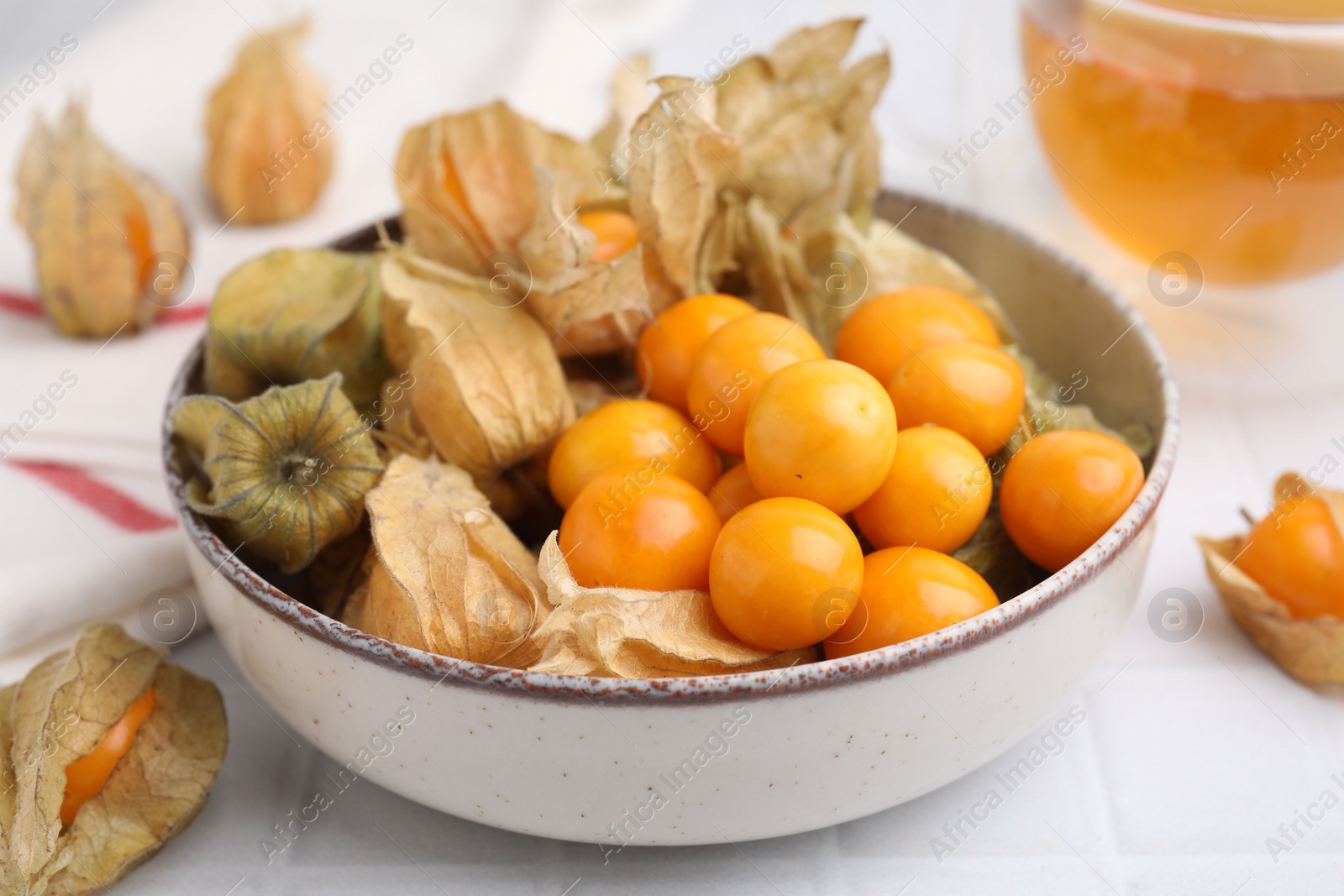 Photo of Ripe physalis fruits with calyxes in bowl on white tiled table, closeup