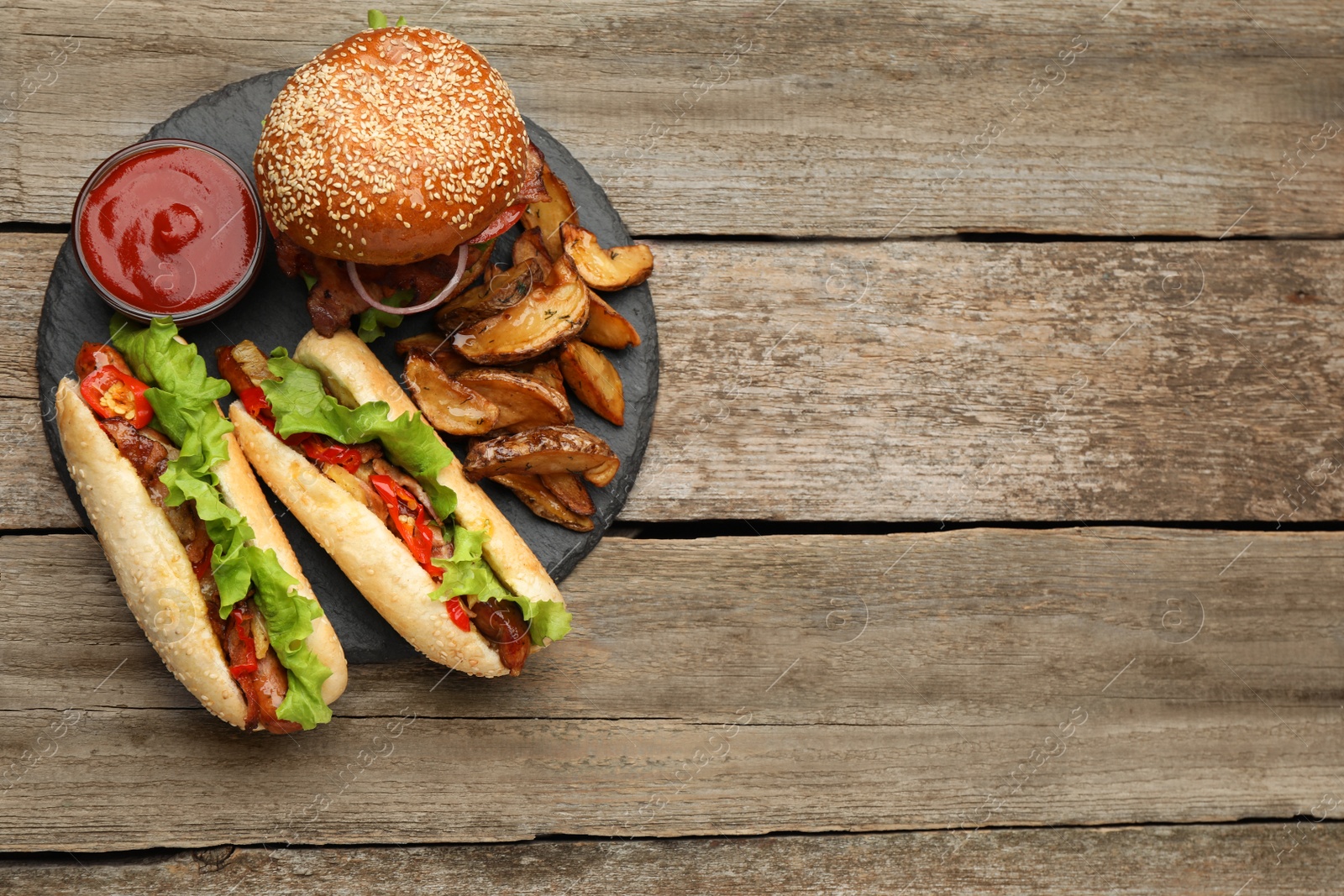 Photo of Tasty burger, hot dogs, potato wedges and sauce on wooden table, top view with space for text. Fast food