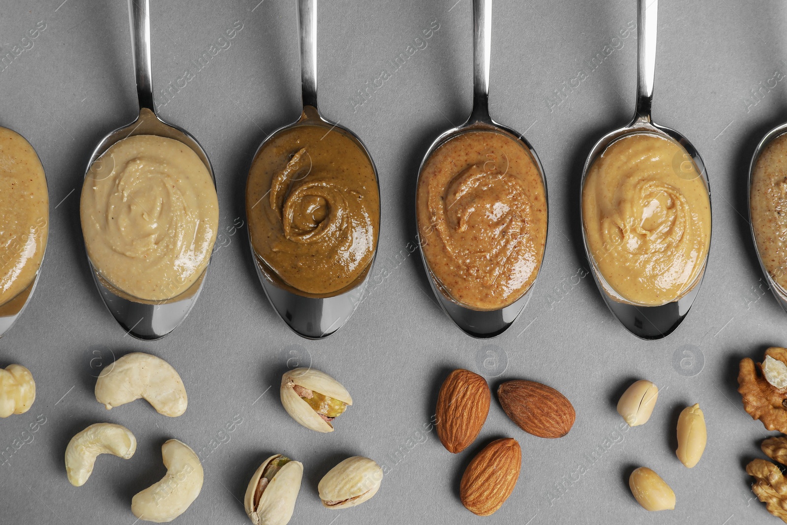 Photo of Tasty nut butters in spoons and raw nuts on gray table, flat lay