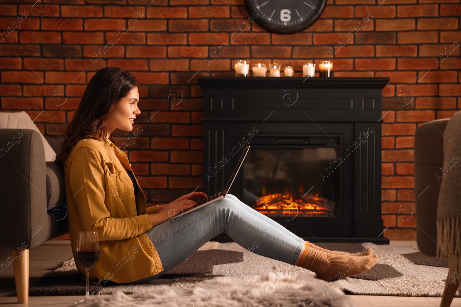 Photo of Young woman with glass of wine and laptop near fireplace at home