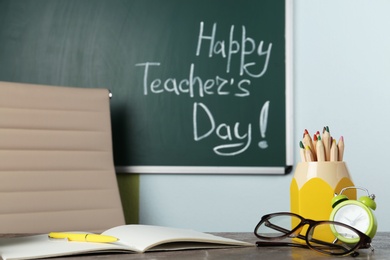 Photo of Eyeglasses and stationery on grey table near chalkboard with inscription HAPPY TEACHER'S DAY in classroom