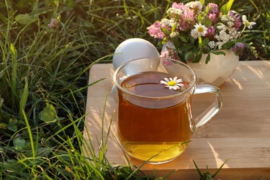 Photo of Cup of aromatic herbal tea, pestle and ceramic mortar with different wildflowers on green grass outdoors