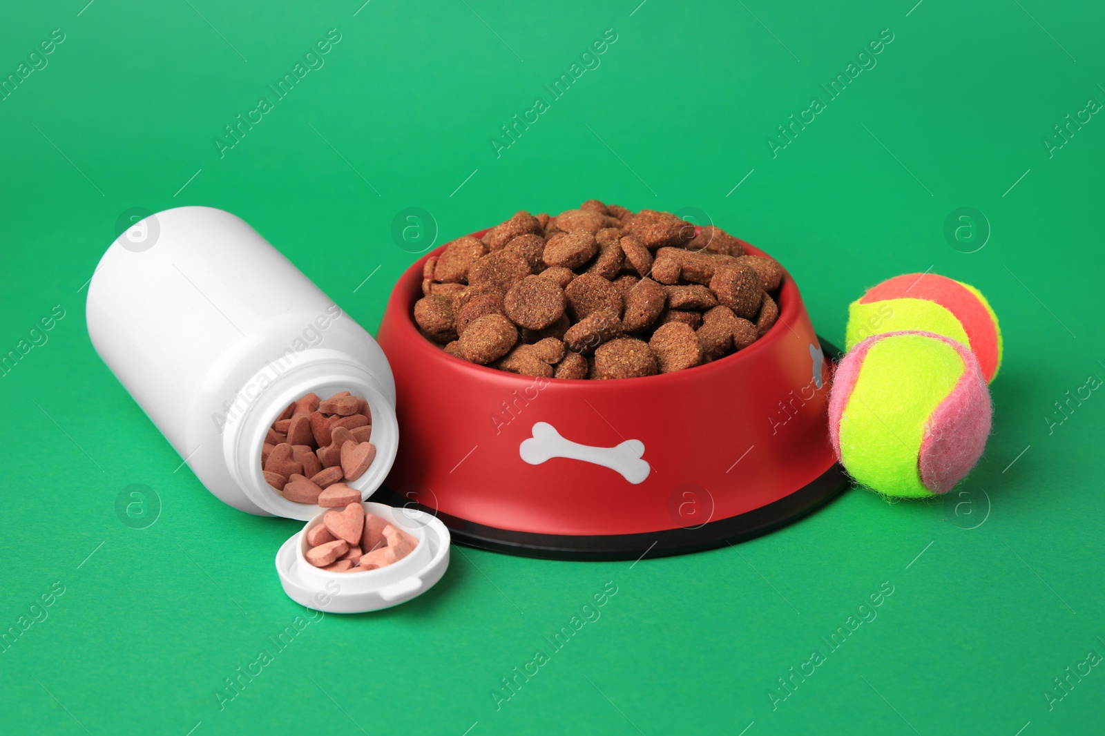 Photo of Bowl with dry pet food, bottle of vitamins and toys on green background