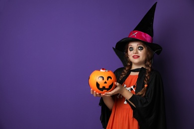 Photo of Cute little girl with pumpkin candy bucket wearing Halloween costume on purple background. Space for text