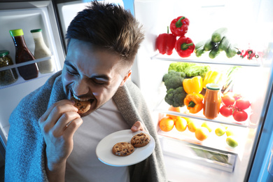Young man eating cookies near open refrigerator at night