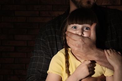 Photo of Adult man covering scared little girl's mouth near brick wall, space for text. Child in danger