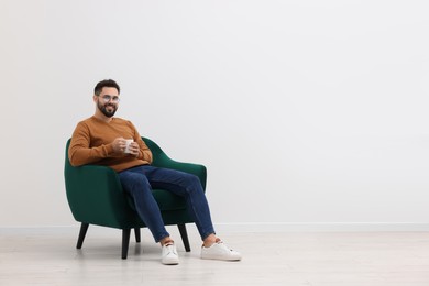 Photo of Handsome man with cup of drink sitting in armchair near white wall indoors, space for text