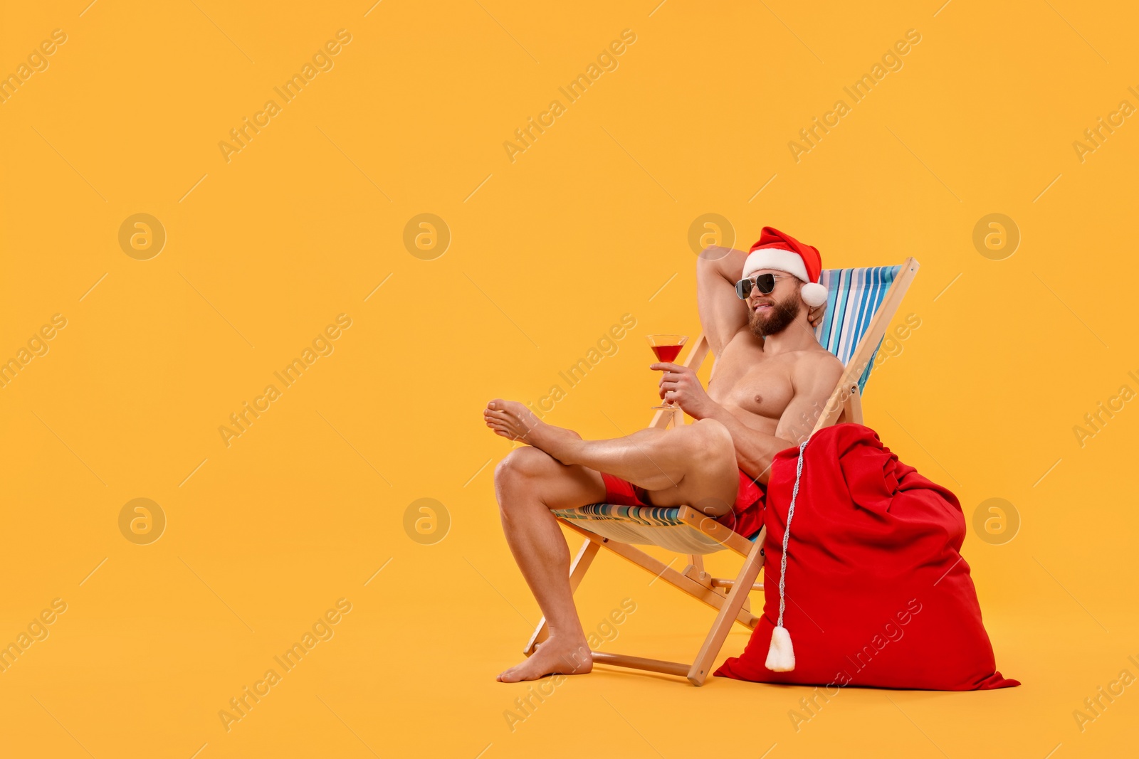 Photo of Muscular young man in Santa hat with deck chair, bag, sunglasses and cocktail on orange background, space for text