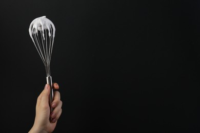 Woman holding whisk with whipped cream on black background, closeup. Space for text