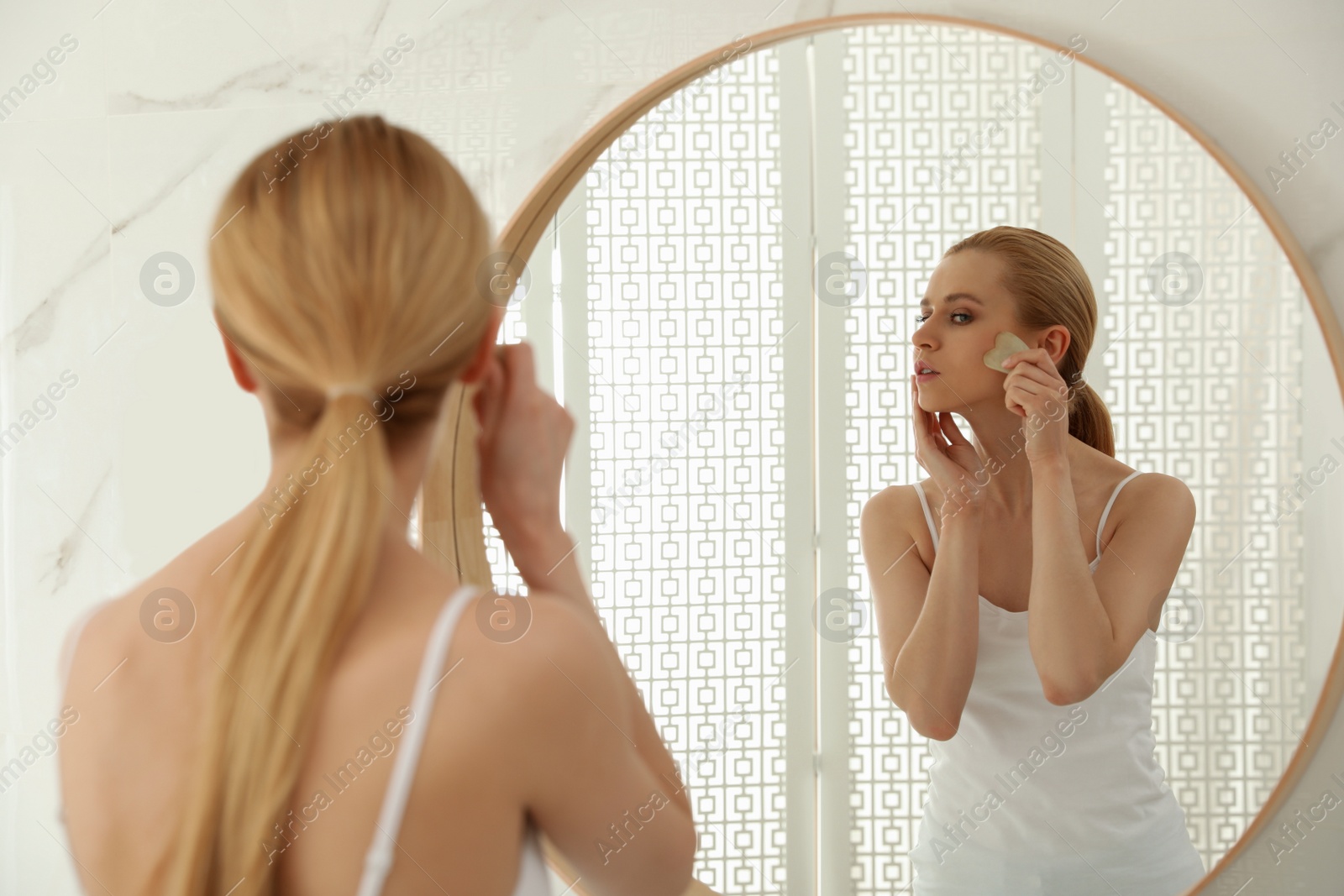 Photo of Beautiful young woman doing facial massage with gua sha tool in front of mirror at home