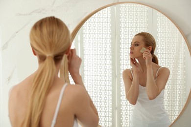 Photo of Beautiful young woman doing facial massage with gua sha tool in front of mirror at home