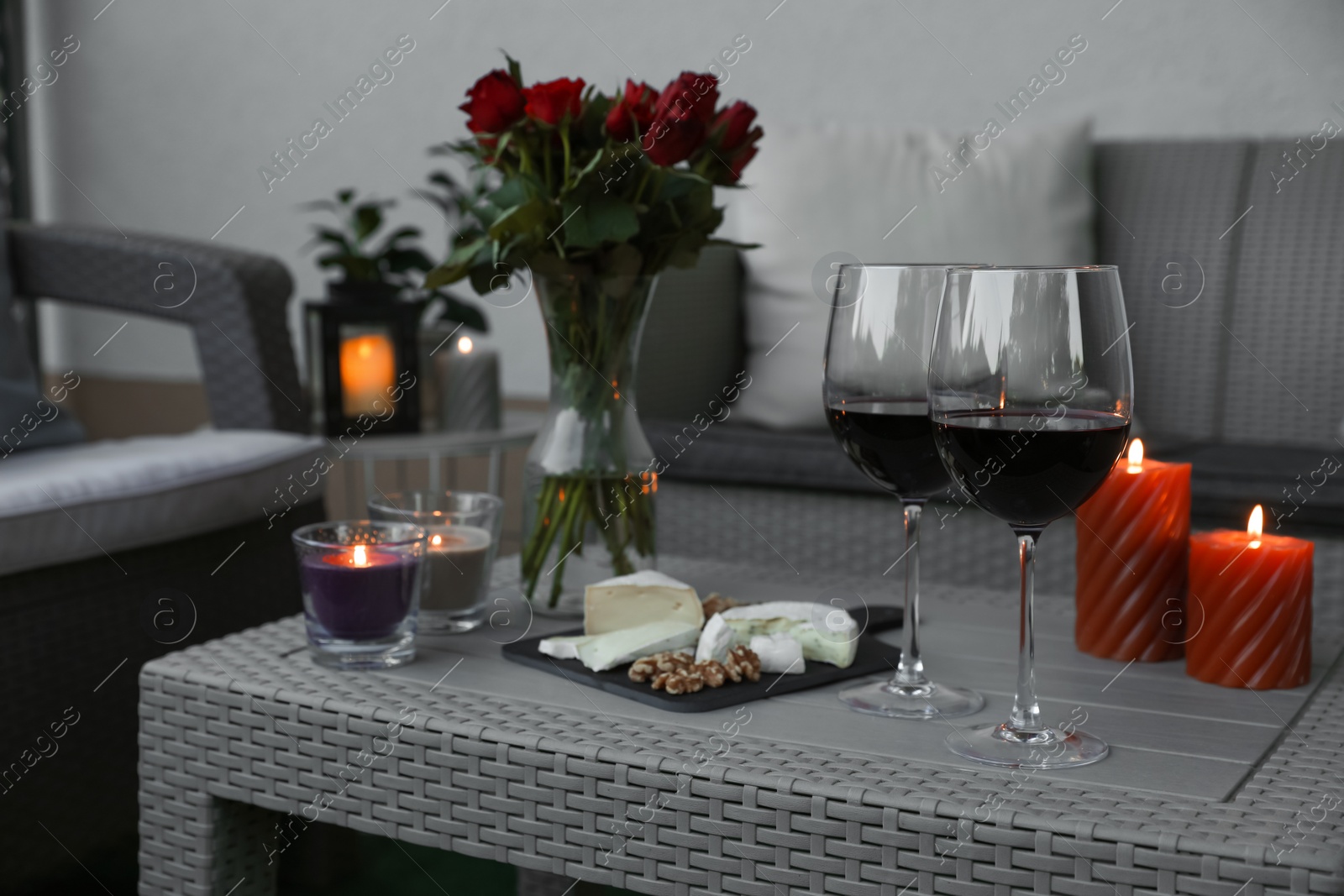 Photo of Glasses of wine, vase with roses, burning candles and snacks on rattan table at balcony in evening, closeup