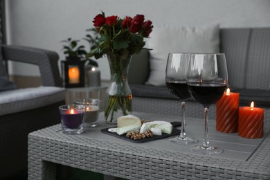 Glasses of wine, vase with roses, burning candles and snacks on rattan table at balcony in evening, closeup