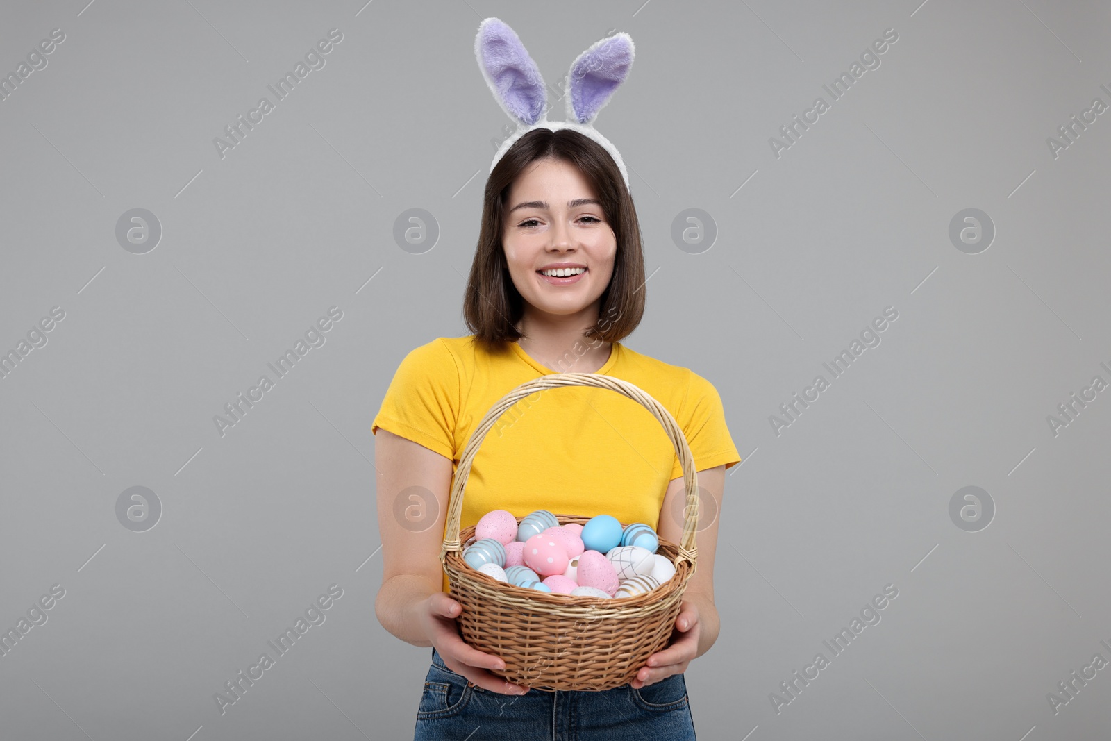 Photo of Easter celebration. Happy woman with bunny ears and wicker basket full of painted eggs on grey background