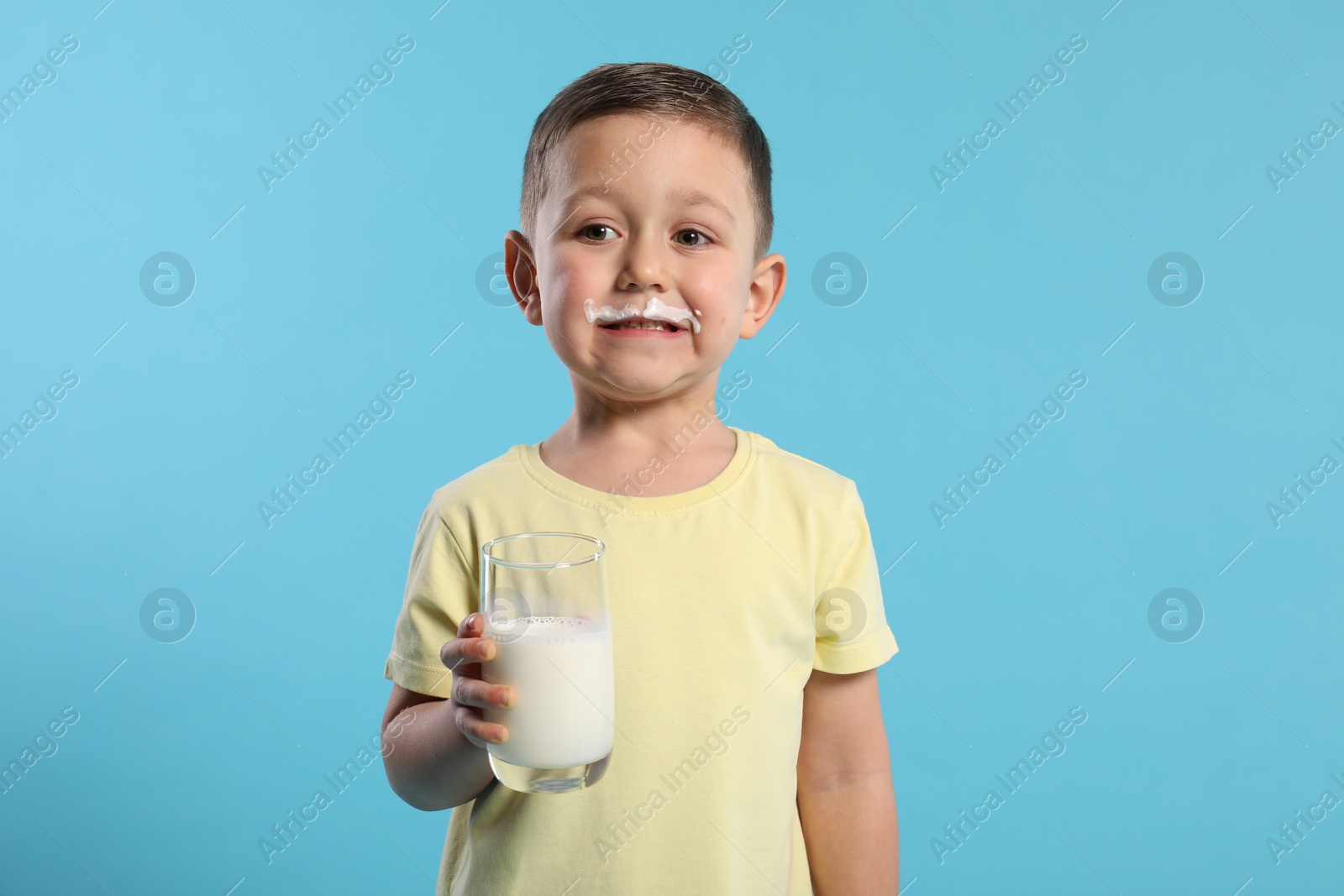 Photo of Cute boy with milk mustache holding glass of tasty dairy drink on light blue background