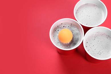 Photo of Plastic cups and ball on red background, above view with space for text. Beer pong game