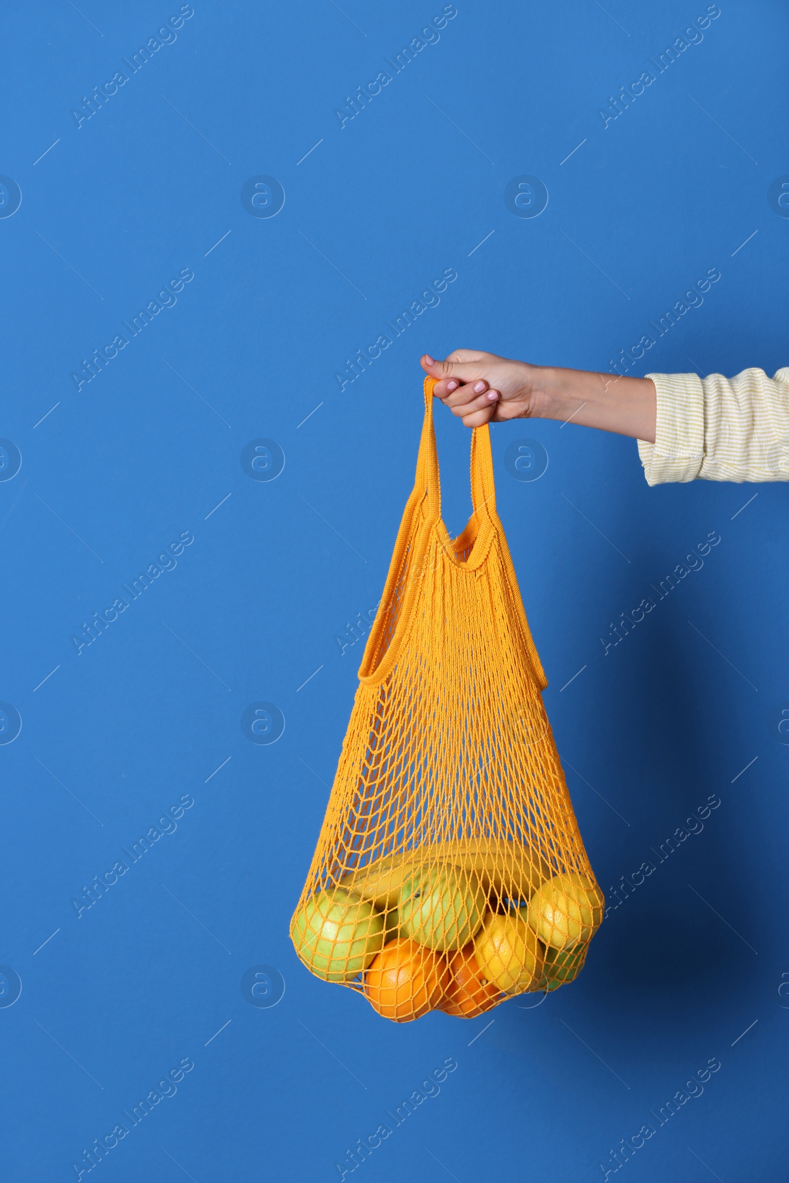 Photo of Woman holding net bag with fruits on blue background, closeup