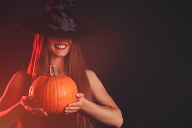 Photo of Young woman wearing witch costume with pumpkin on dark background, space for text. Halloween party