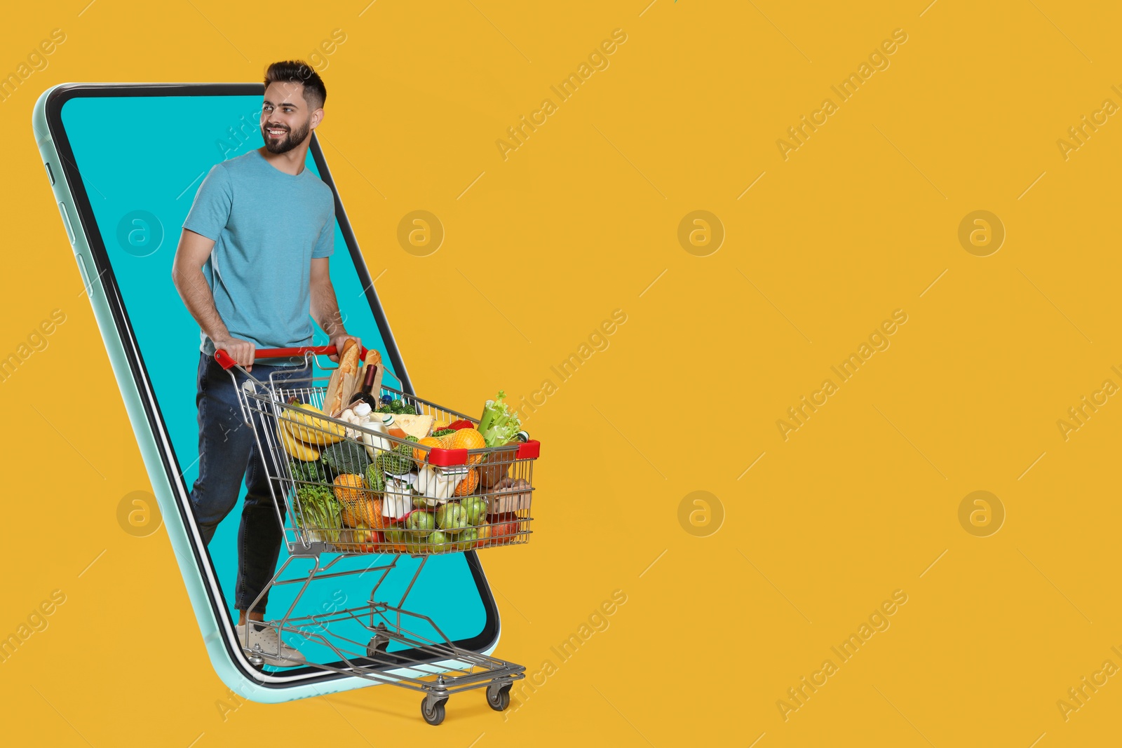 Image of Grocery shopping via internet. Happy man with shopping cart full of products walking out of huge smartphone on orange background, space for text