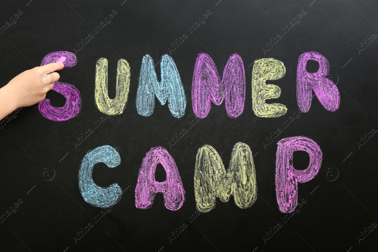 Photo of Child writing SUMMER CAMP with colorful chalk on blackboard, closeup