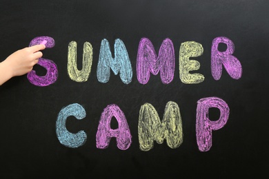 Child writing SUMMER CAMP with colorful chalk on blackboard, closeup