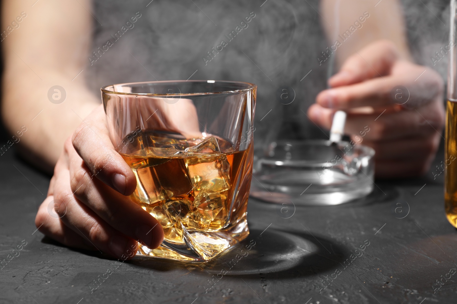 Photo of Alcohol addiction. Man with glass of whiskey and smoldering cigarette at dark textured table, selective focus