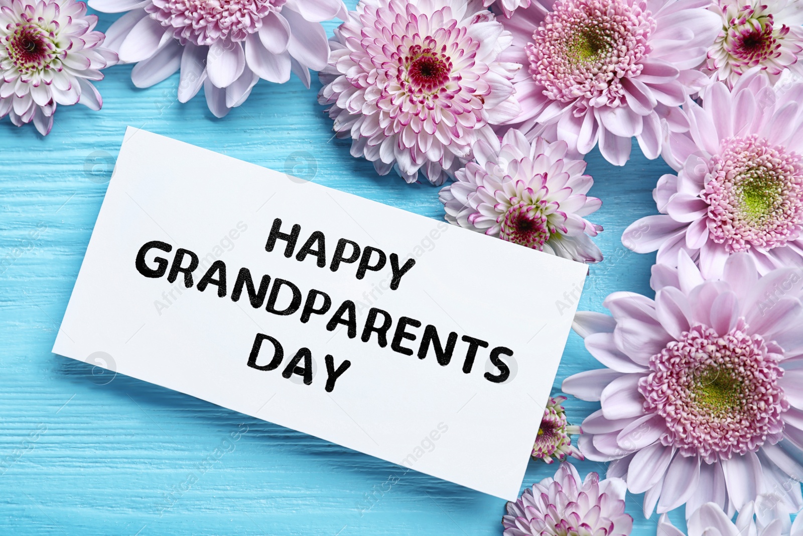 Photo of Beautiful flowers and card with phrase Happy Grandparents Day on light blue wooden background, flat lay