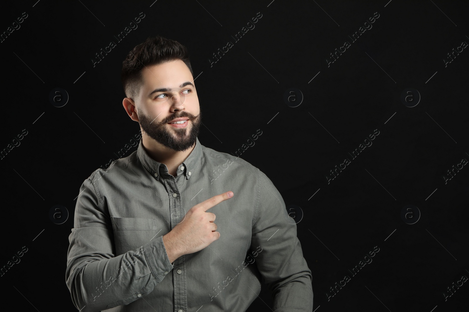 Photo of Young man with mustache pointing at something on black background. Space for text