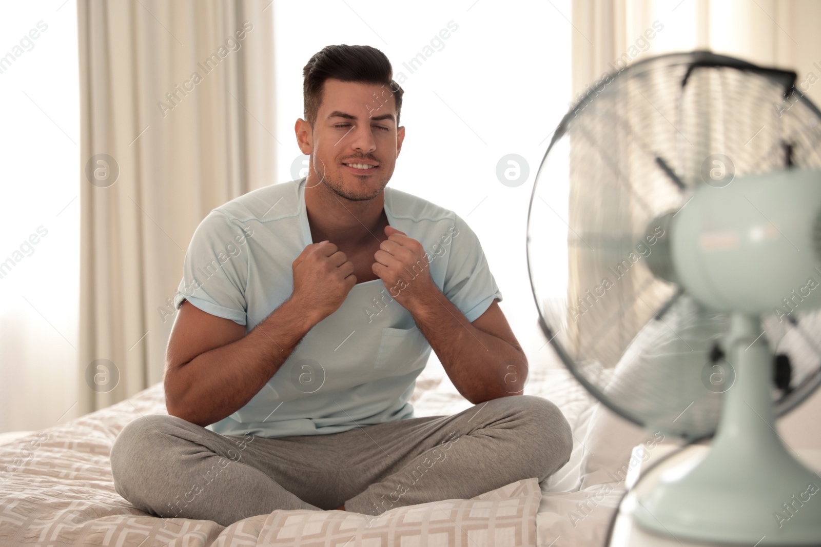 Photo of Man enjoying air flow from fan on bed in room. Summer heat