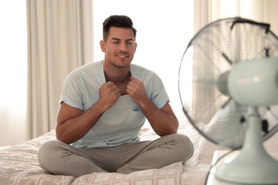 Man enjoying air flow from fan on bed in room. Summer heat