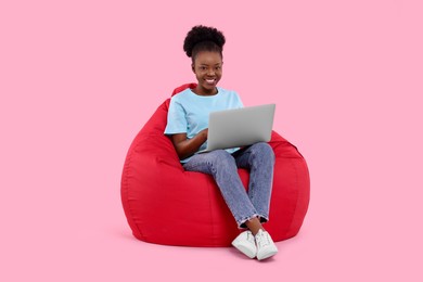 Beautiful young woman with laptop sitting on beanbag chair against pink background