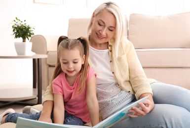 Photo of Mature woman with her little granddaughter reading book together at home