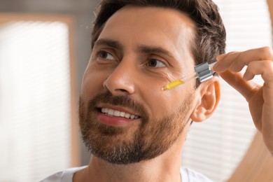 Photo of Handsome man applying cosmetic serum onto his face indoors, closeup