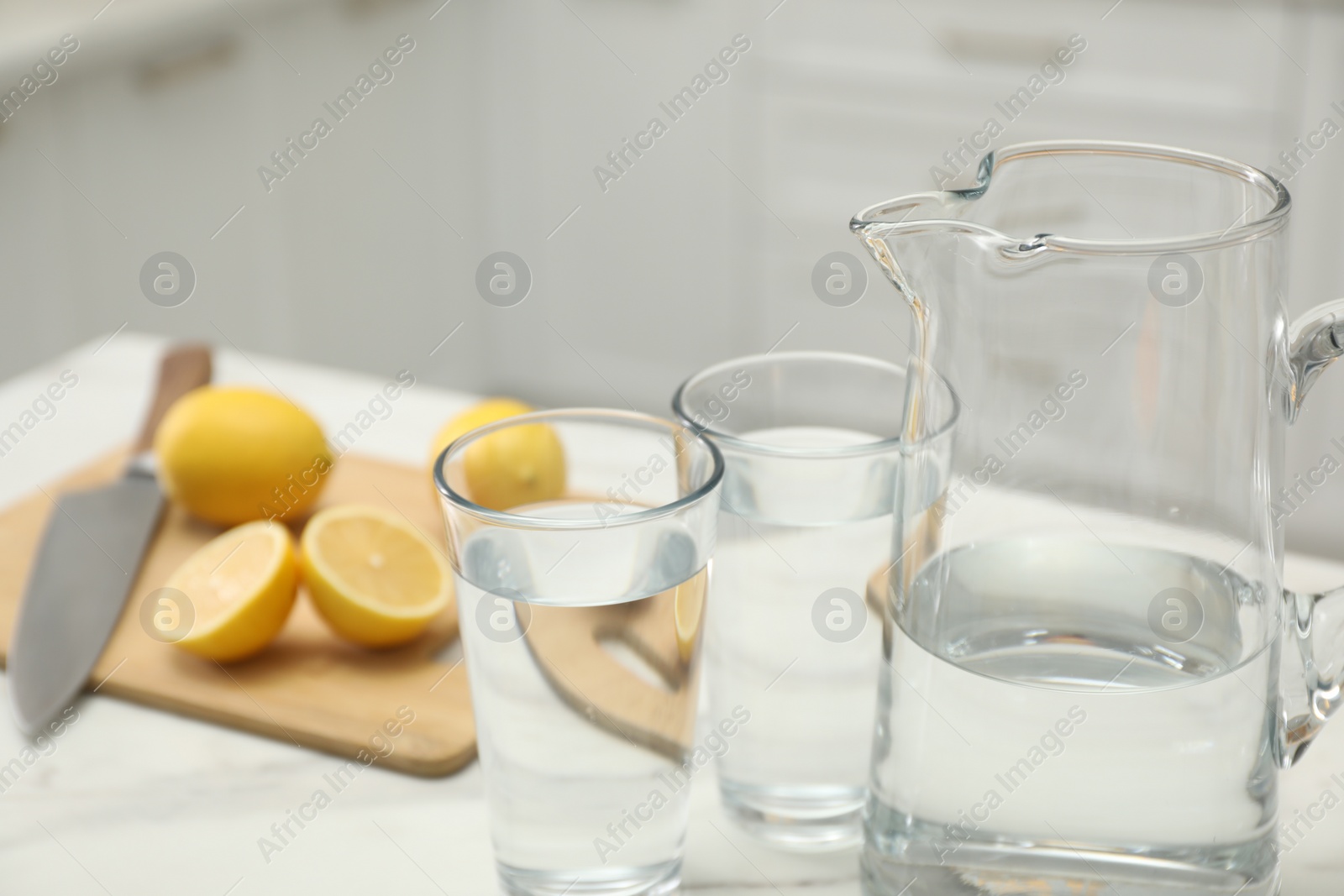 Photo of Jug, glasses with clear water and lemons on white table in kitchen, closeup