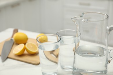 Jug, glasses with clear water and lemons on white table in kitchen, closeup