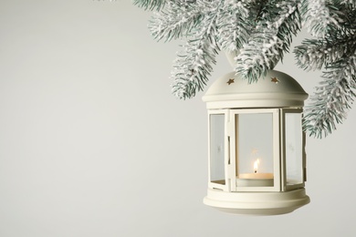 Photo of Christmas lantern with candle hanging on snowy fir tree branch against light background. Space for text