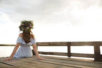 Photo of Young woman wearing wreath made of beautiful flowers on pier near river, back view