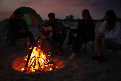 Photo of Group of friends gathering around bonfire on beach in evening. Camping season