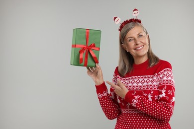 Happy senior woman in Christmas sweater and Santa headband holding gift on grey background. Space for text