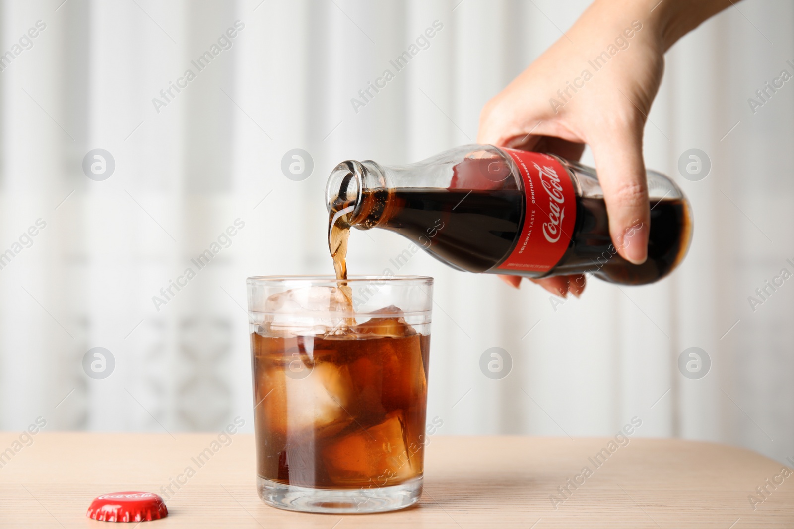 Photo of MYKOLAIV, UKRAINE - NOVEMBER 15, 2018: Woman pouring Coca Cola into glass with ice cubes at table, closeup