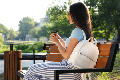 Photo of Beautiful young woman with stylish backpack and cup of coffee on bench in park