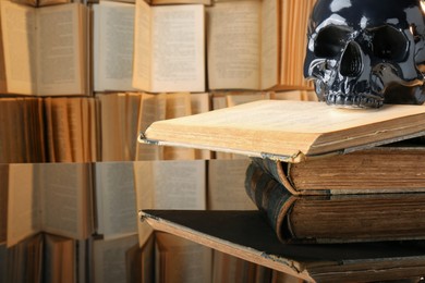 Photo of Black human skull and old books on mirror table. Space for text