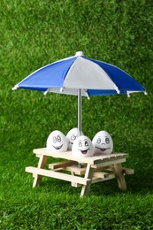Photo of Eggs with drawn happy faces, small picnic table and umbrella on green grass