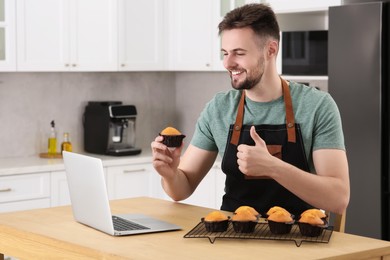 Photo of Man holding muffin near laptop and showing thumb up at table in kitchen. Time for hobby