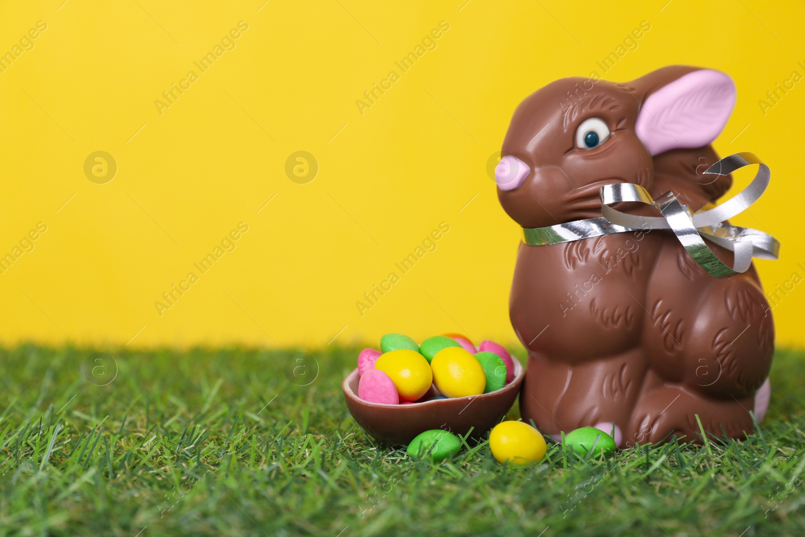 Photo of Easter celebration. Cute chocolate bunny and half of sweet egg with small candies on grass against yellow background. Space for text
