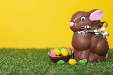 Easter celebration. Cute chocolate bunny and half of sweet egg with small candies on grass against yellow background. Space for text