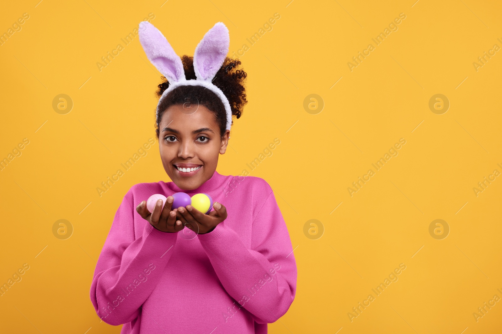 Photo of Happy African American woman in bunny ears headband holding Easter eggs on orange background, space for text